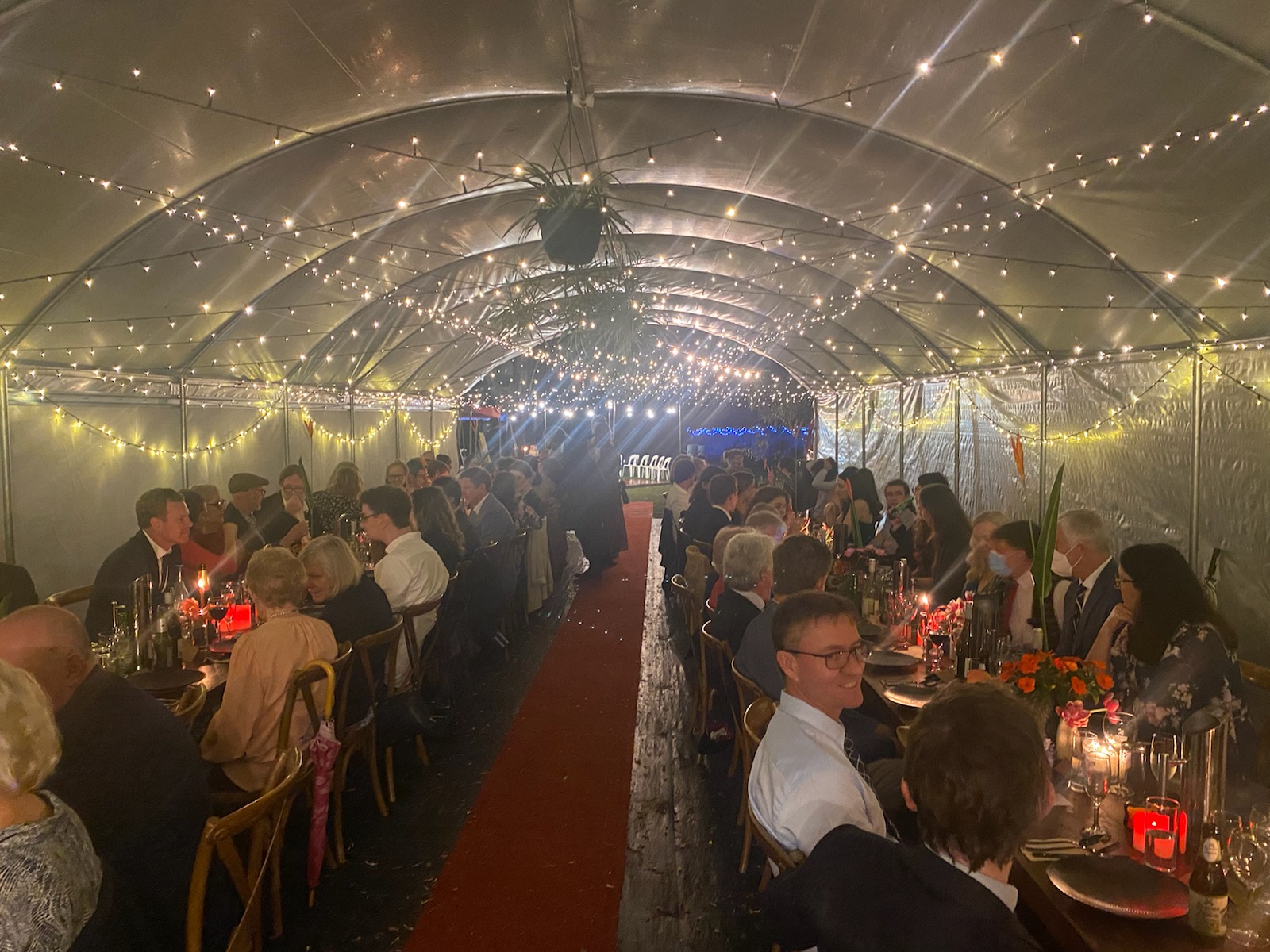 Group of people celebrating together, sitting on two rows of long tables, center red carpet, fairy lights on ceiling