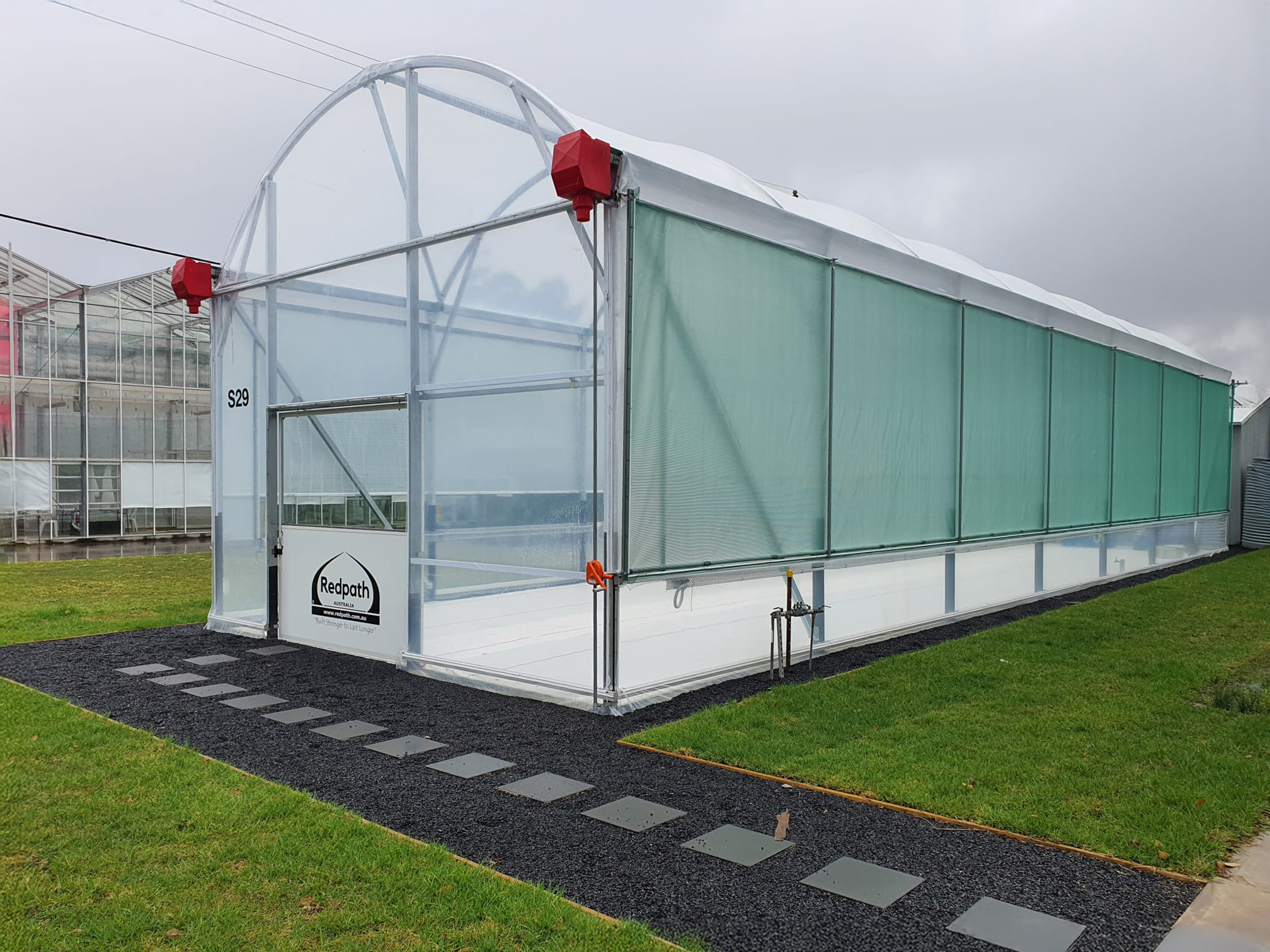 Exterior shot of medium sized arch greenhouse - redpath branding on front - surrounded by grass with stone tiles path leading to entrance of greenhouse